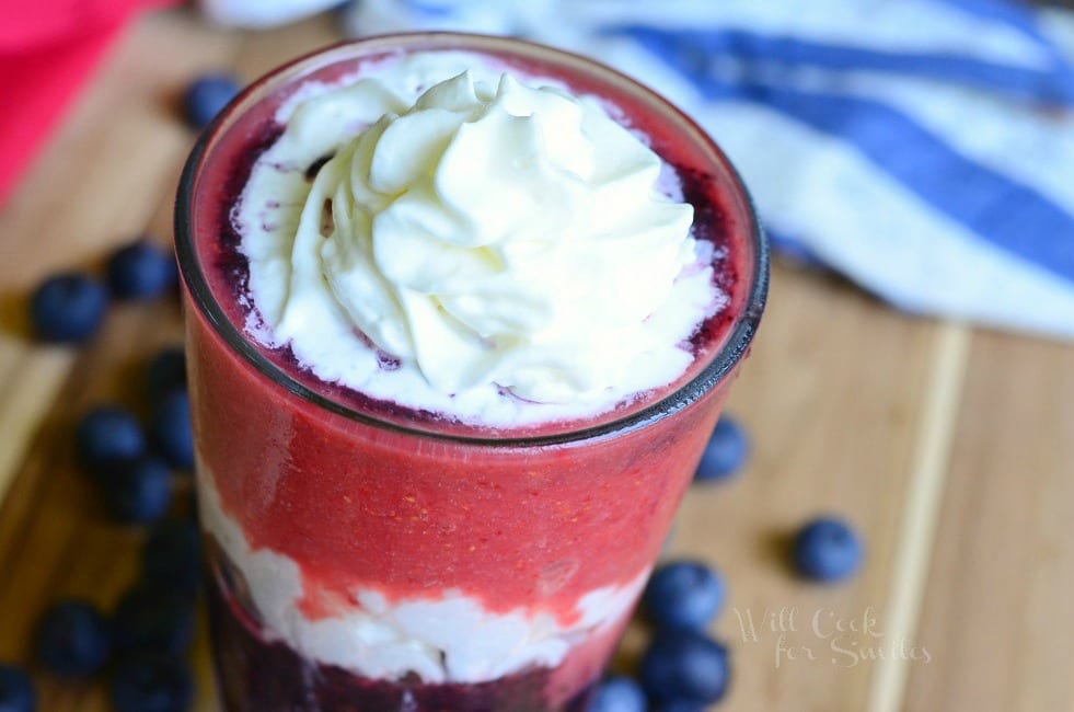 Layered Berry Daiquiri Red White and Blue Daiquiri is in a tall glass. There are layers of whipped cream and berries up to the rim. The top has whipped cream as well. Blueberries are scattered around the glass on a wooden board.
