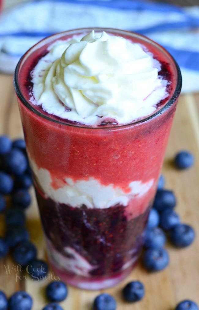 Layered Berry Daiquiri Red White and Blue Daiquiri is in a tall glass. There are layers of whipped cream and berries up to the rim. The top has whipped cream as well. Blueberries are scattered around the glass on a wooden board.