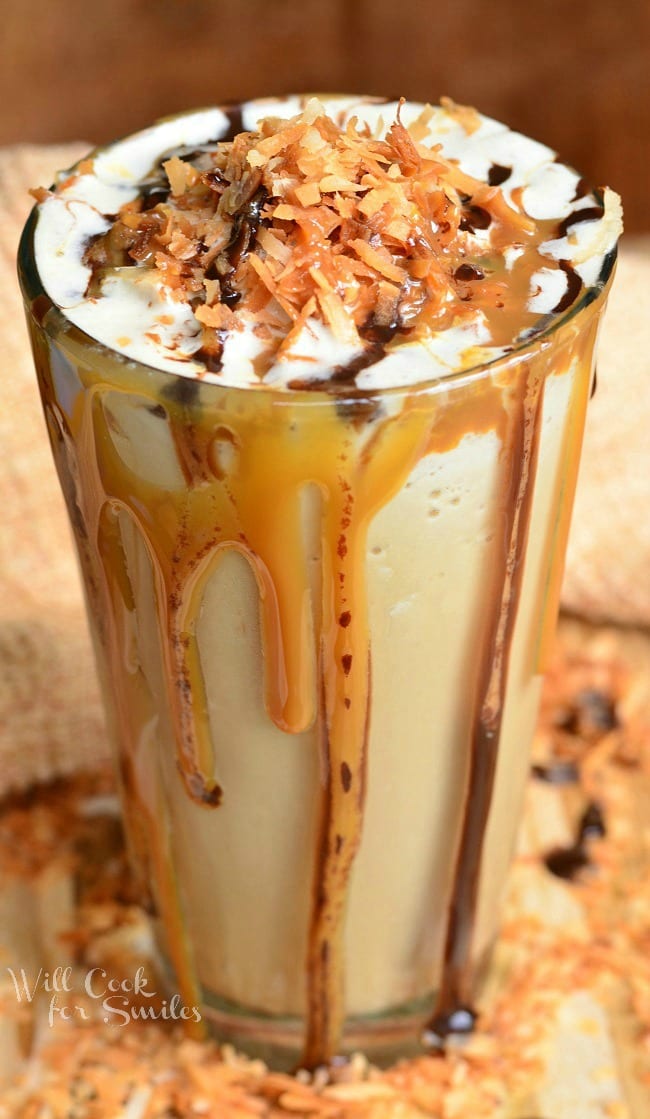 Samoas Frozen Coffee in a glass cup with whip cream, toasted coconut, chocolate sauce, and caramel on top. Caramel and chocolate sauce pouring down the outside of the glass 