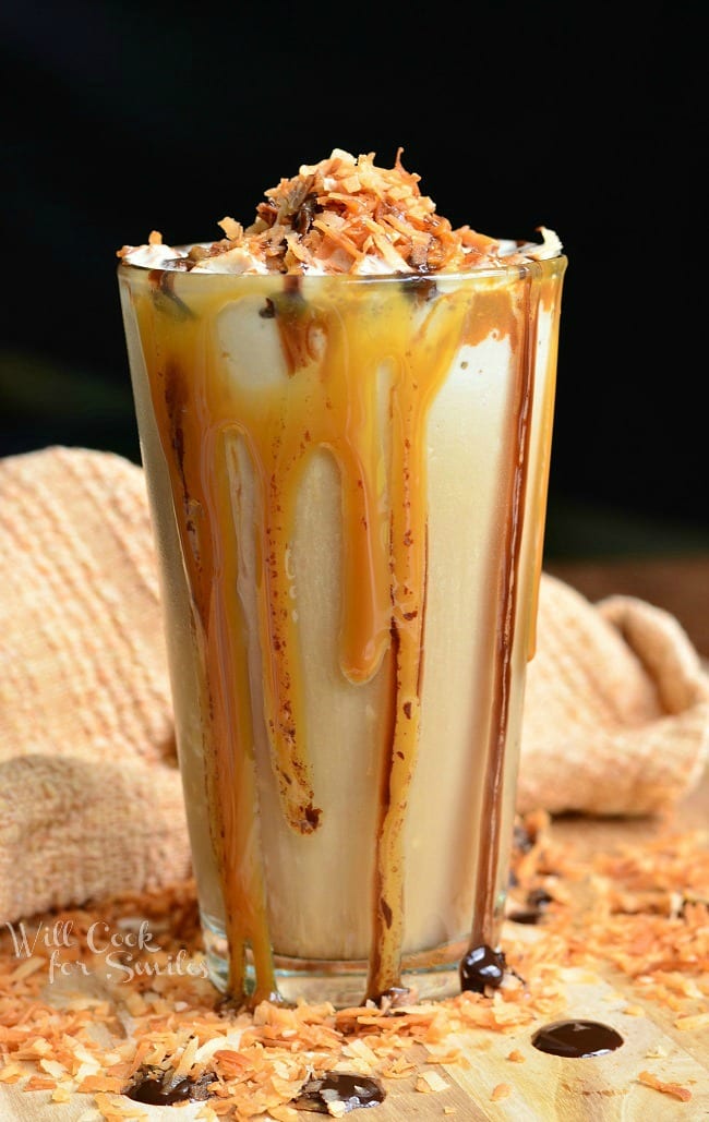Samoas Frozen Coffee in a glass cup with whip cream, toasted coconut, chocolate sauce, and caramel on top. Caramel and chocolate sauce pouring down the outside of the glass 