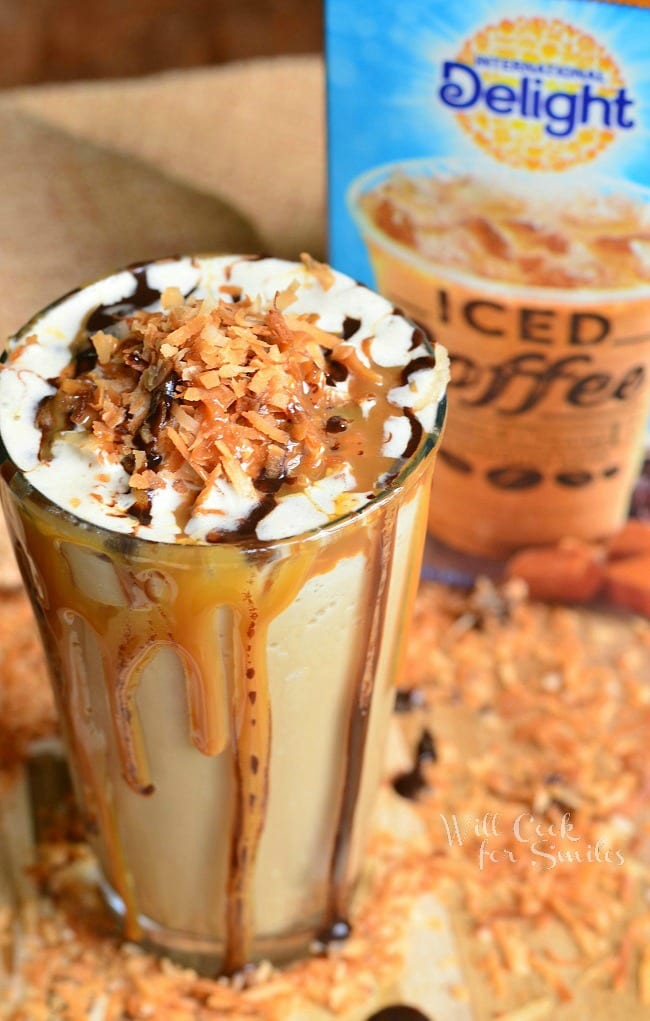 Samoas Frozen Coffee in a glass cup with whip cream, toasted coconut, chocolate sauce, and caramel on top. Caramel and chocolate sauce pouring down the outside of the glass with a carton of delight iced coffee in the background
