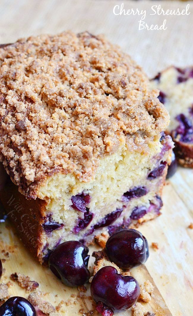 A few pieces of Cherry Streusel Bread have been cut from the loaf. This photo is of the remainder of the loaf. The streusel is on top of the bread and laying around the wooden board. There are also some fresh cherries laying there as well.