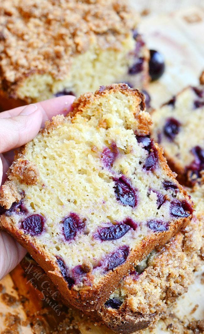 A few pieces of Cherry Streusel Bread have been cut from the loaf. A hand holds one piece of the bread. The streusel is on top of the bread and laying around the bread slices.