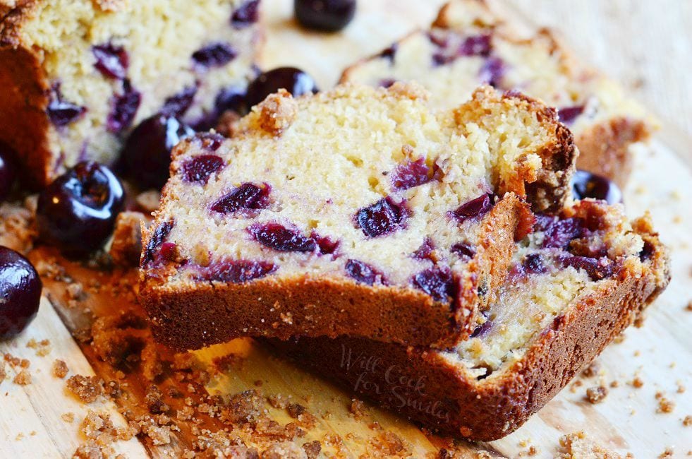 A few pieces of Cherry Streusel Bread have been cut from the loaf. The cut pieces lay stacked on each other in front of the loaf. The streusel is on top of the bread and laying around the bread slices. There's also fresh cherries on the wooden surface.