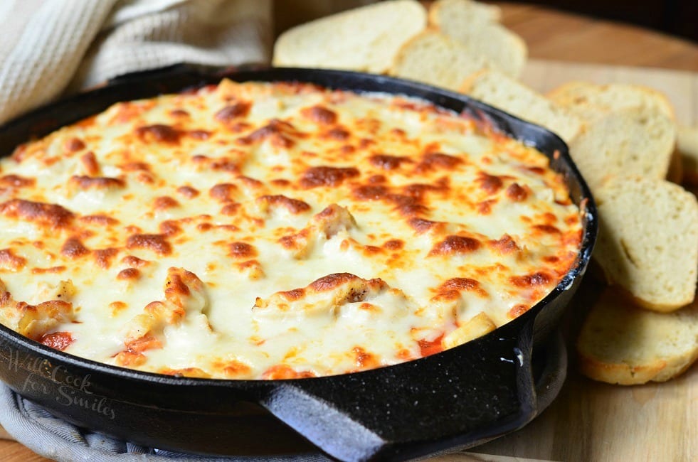 Chicken Parmesan Dip in a black skillet. The top is cheese that has been browned to perfection. Slices of bread are behind the skillet.