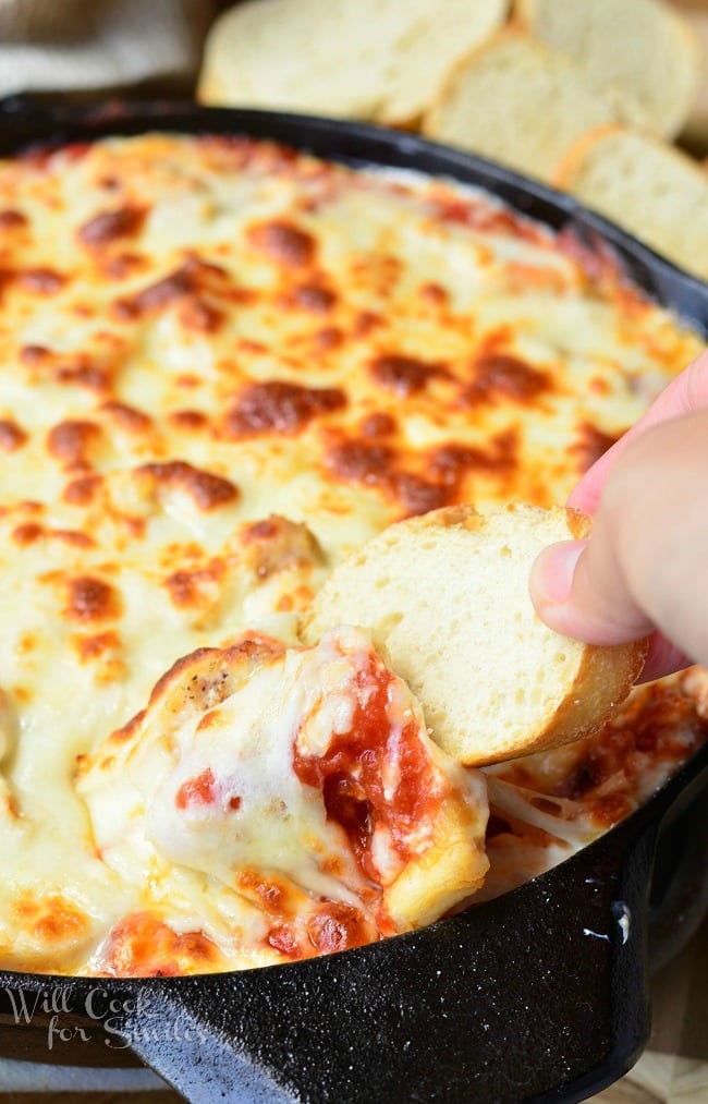 Chicken Parmesan Dip in a black skillet. The top is cheese that has been browned to perfection. A right hand is taking a piece of bread to dip into the skillet to scoop some of the dip up. The cheese looks gooey.