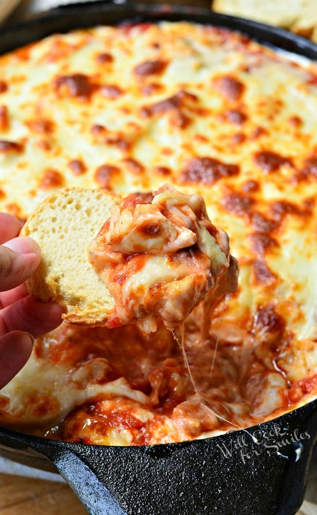 Chicken Parmesan Dip in a black skillet. The top is cheese that has been browned to perfection. A left hand is holding a piece of bread over the skillet that has the gooey dip on top. The cheese is stringy.