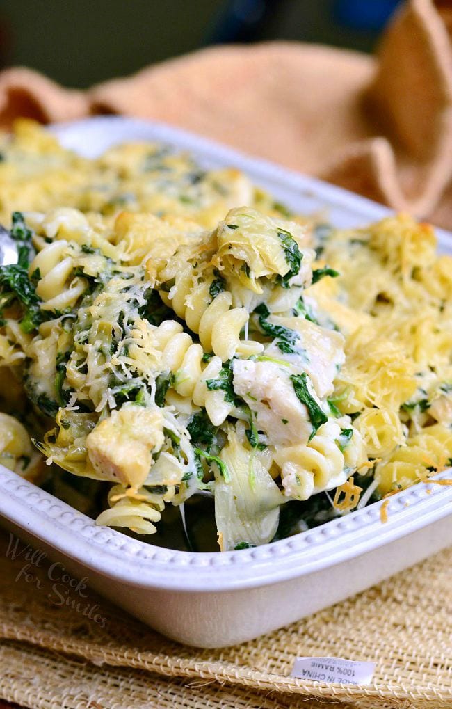 Spoon lifting up some of the Chicken Spinach and Artichoke Pasta that is served in a white casserole dish. 