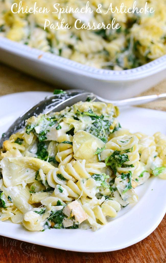 A serving of the Chicken Spinach and Artichoke Pasta Casserole on a white plate with serving spoon. White casserole dish with pasta fades into the background.
