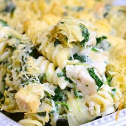 white baking dish filled with chicken spinach and artichoke pasta casserole on a tan placemat with a brown napkin in the background