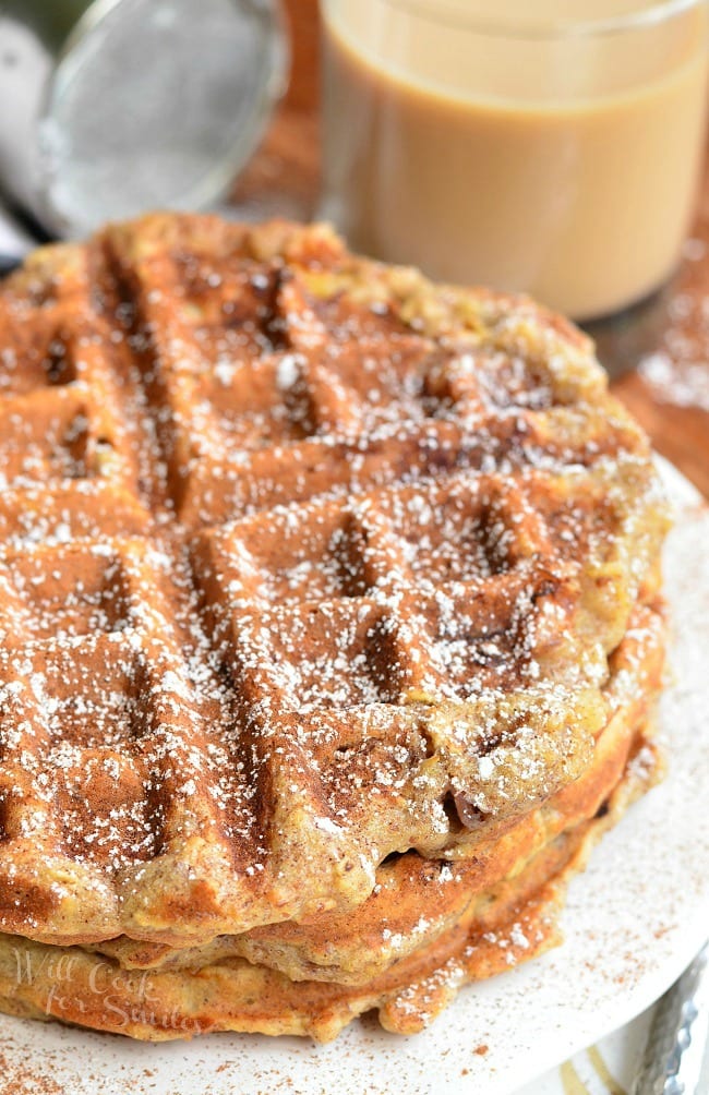 Top view of three Cinnamon Peach Oat Waffles stacked on top of each other served on a white plate. Waffles are sprinkled with cinnamon and powder sugar.