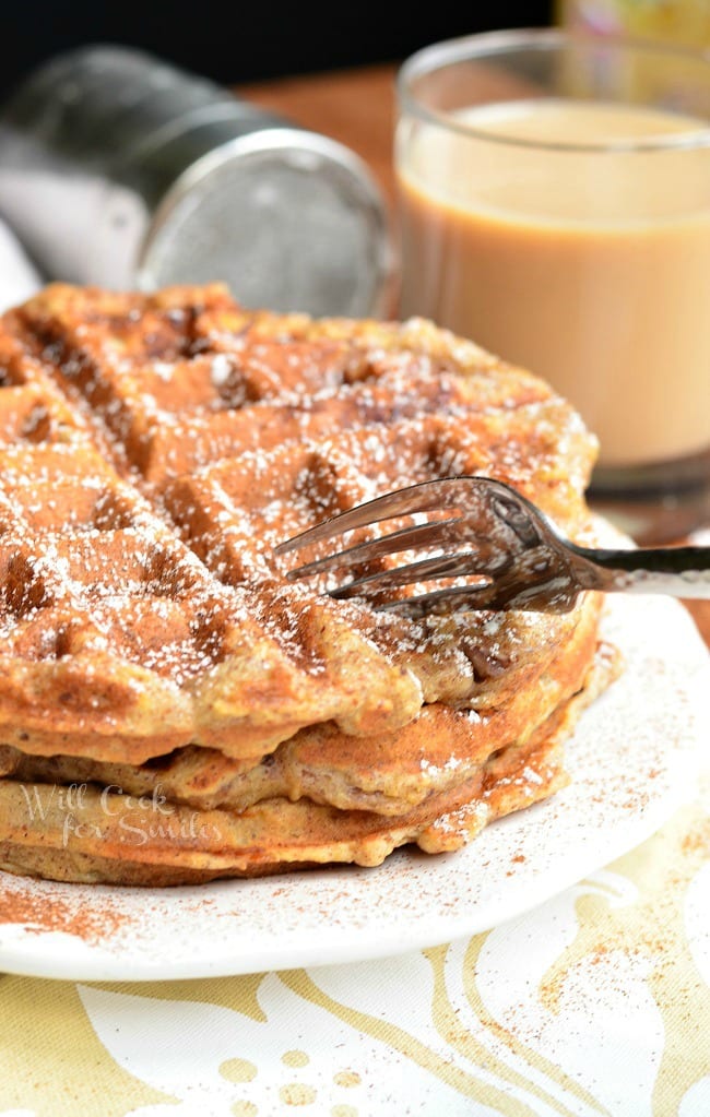 Three Cinnamon Peach Oat Waffles stacked on top of each other served on a white plate. Waffles are sprinkled with cinnamon and powder sugar. A fork is inserted into the top of the waffle stack.