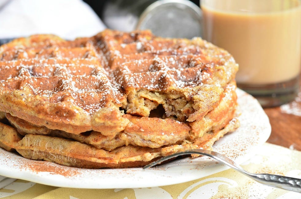 Side view of three Cinnamon Peach Oat Waffles stacked on top of each other served on a white plate. Waffles are sprinkled with cinnamon and powder sugar. A bite or piece of the top waffle is gone and the fork is set on the edge of the plate.