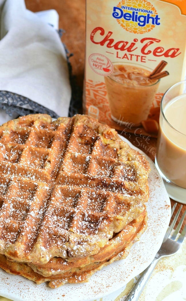 Top view of three Cinnamon Peach Oat Waffles stacked on top of each other served on a white plate. Waffles are sprinkled with cinnamon and powder sugar. A fork is laying on the table to the right of the waffles. A container and glass of Chai Tea Latte is in the back.