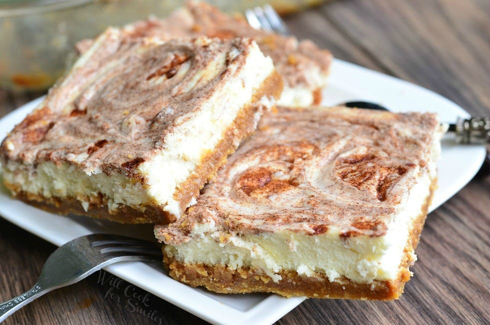Several Caramel Cinnamon Swirl Cheesecake Bars lay piled on a small, white plate. A fork sets on the left rim of the plate.