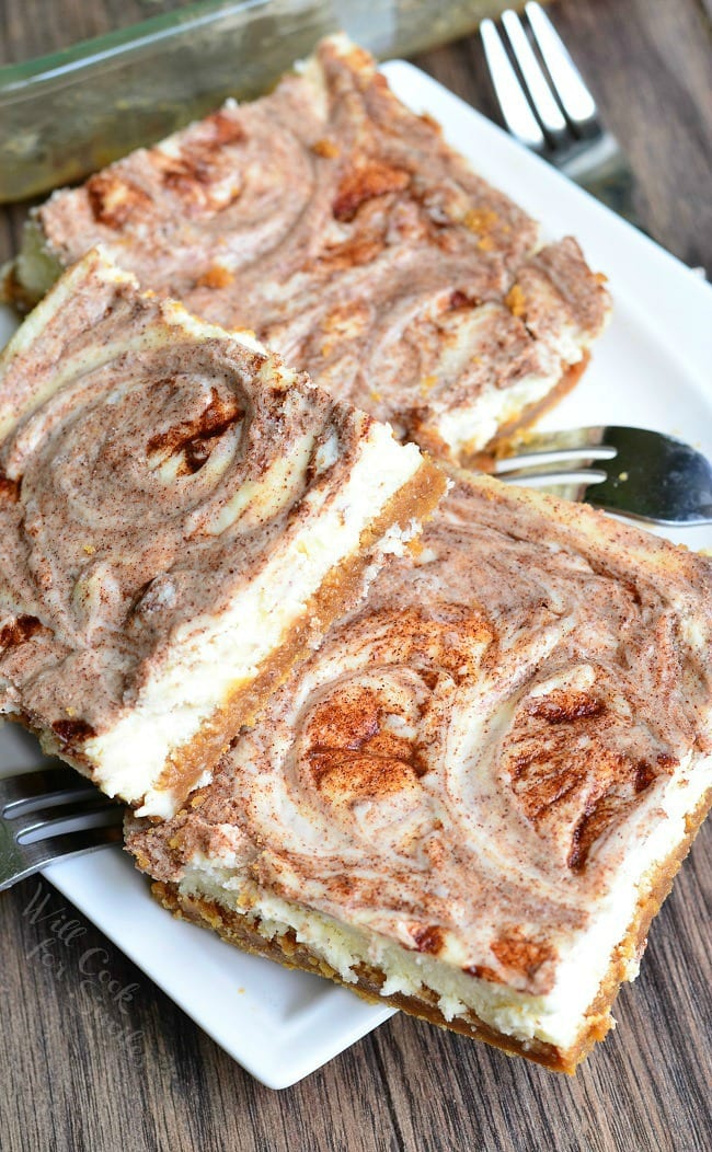 Several Caramel Cinnamon Swirl Cheesecake Bars lay piled on a small, white plate. Several forks also lay on the plate as well.