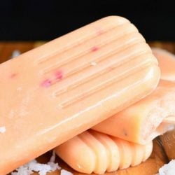 close up view of 3 coconut and peach creamy popsicles on a wooden table with coconut shaving scattered in front of popsicles