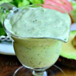 Sauce boat filled with creamy avocado dressing on a wood table with a salad on a white plate in the background