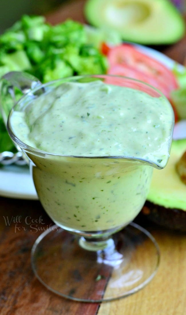 More of a top view of Creamy Avocado Dressing in a glass pouring dish. Fresh vegetables are on a white plate behind the dressing.