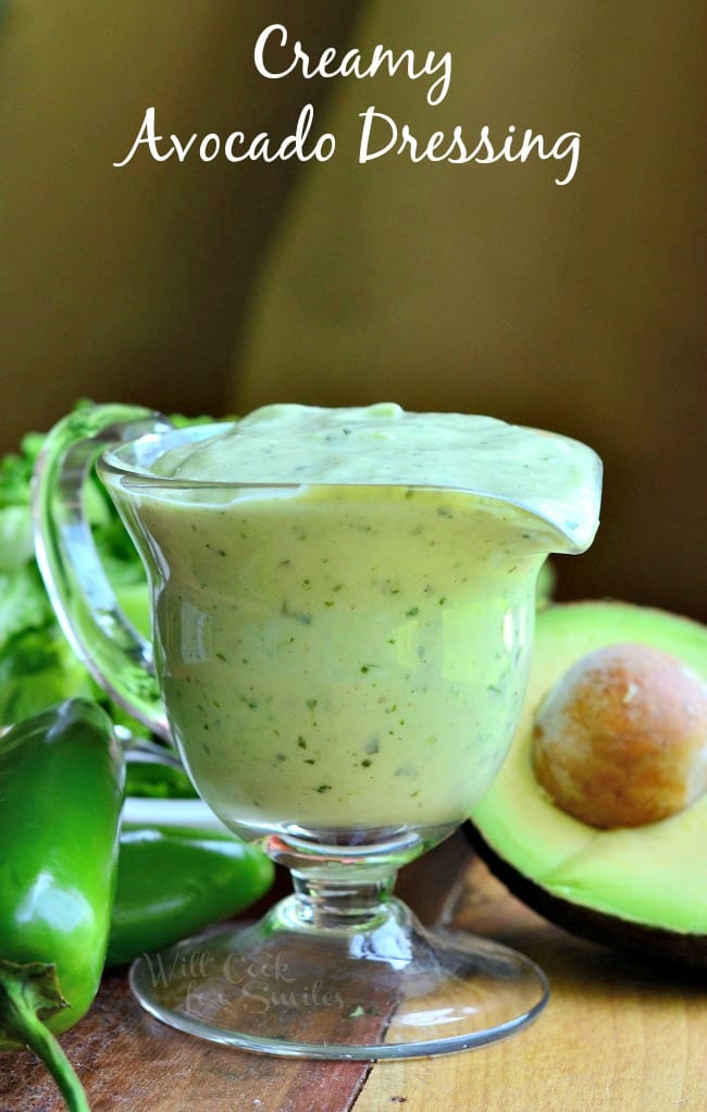 Creamy Avocado Dressing is presented in a glass pouring dish. Green jalapenos and a halved avocado lay on the table beside it.