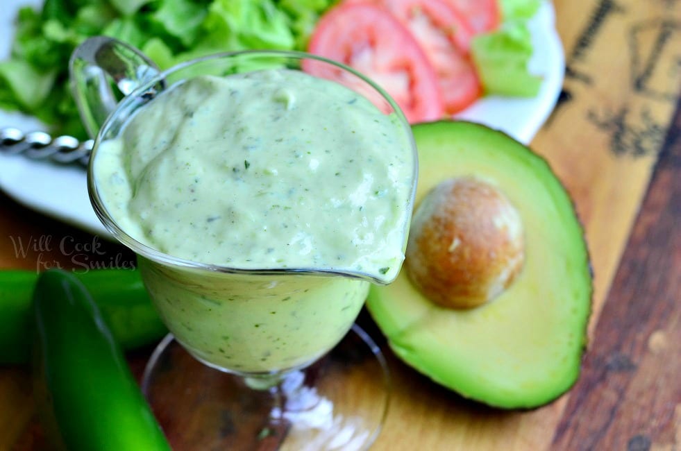 Top view of Creamy Avocado Dressing in a glass pouring dish. Green jalapenos and a halved avocado lay on the table beside it. A plate of vegetables behind it.