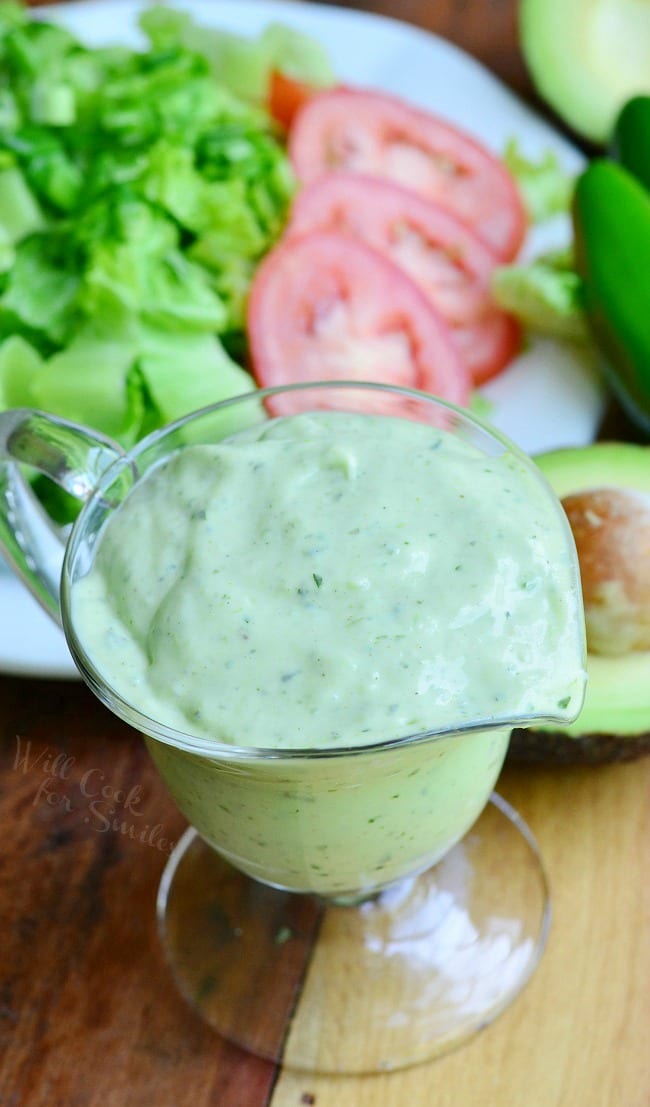 Top view of Creamy Avocado Dressing in a glass pouring dish. Green jalapenos and a halved avocado lay on the table behind it, as well as a plate of lettuce and slice tomato.