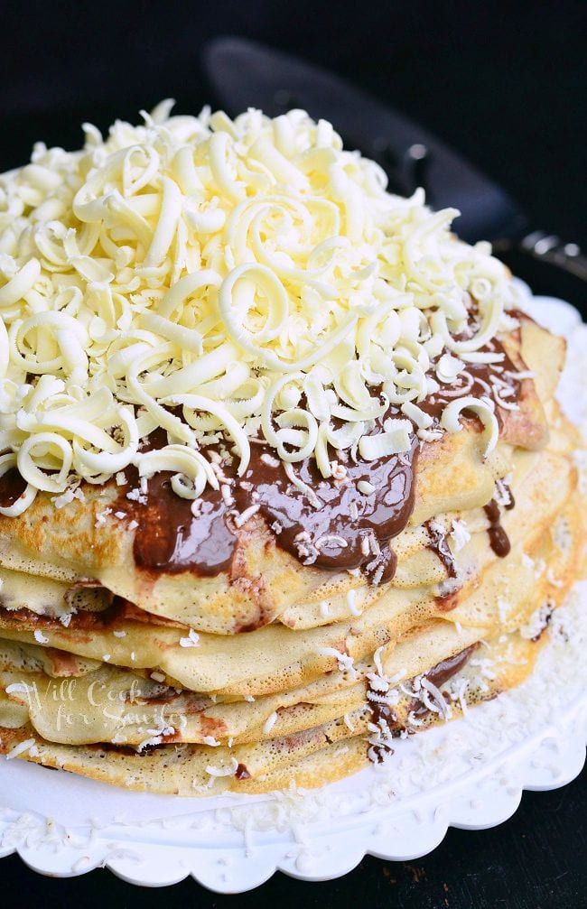 Double Chocolate Chocolate Pudding Crepe Cake is served on a white dish. The crepes are stacked on one another with filling between layers. The cake is topped with more chocolate pudding and white chocolate shavings.