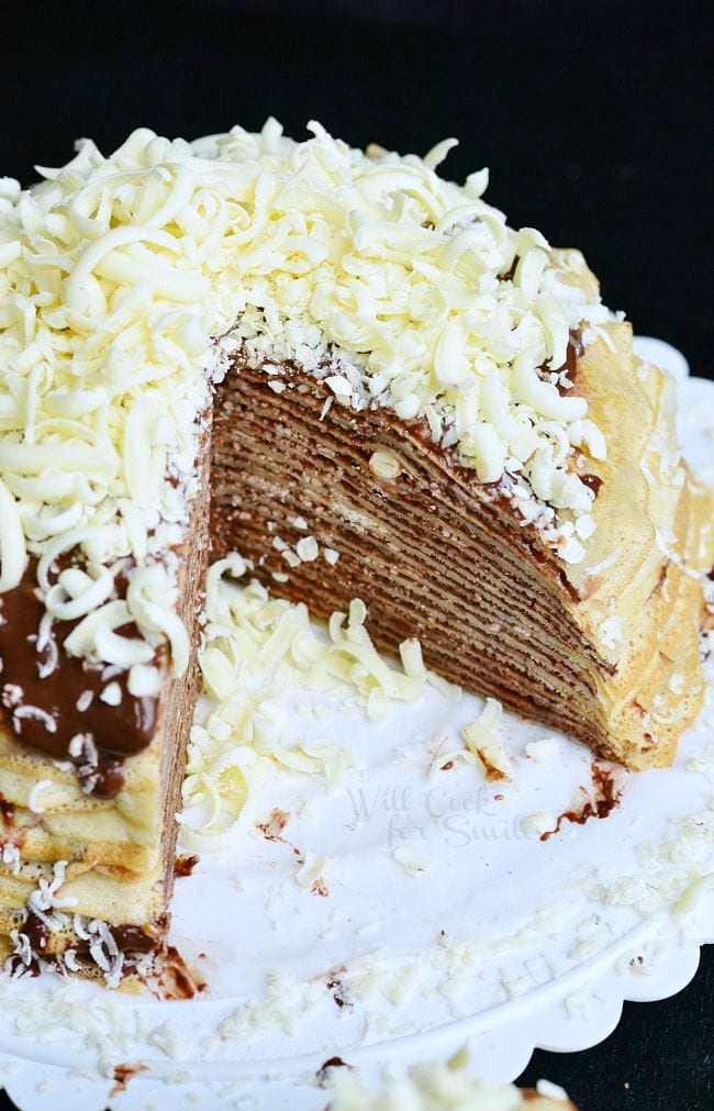 Double Chocolate Chocolate Pudding Crepe Cake is served on a white dish. The crepes are stacked on one another with filling between layers. The cake is topped with more chocolate pudding and white chocolate shavings. One piece of the cake is missing allowing the inside layers to be seen.