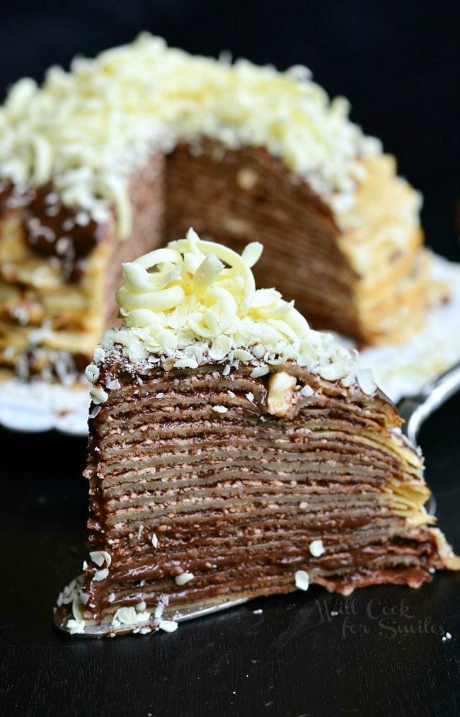 A slice of the Double Chocolate Chocolate Pudding Crepe Cake sits on a serving spatula. The stacked crepes make for thin layers. Chocolate is a key element of this cake. It's between all layers and then on top as well. There are also white chocolate shavings on top of this slice of cake. The rest of the cake fades into the background. 