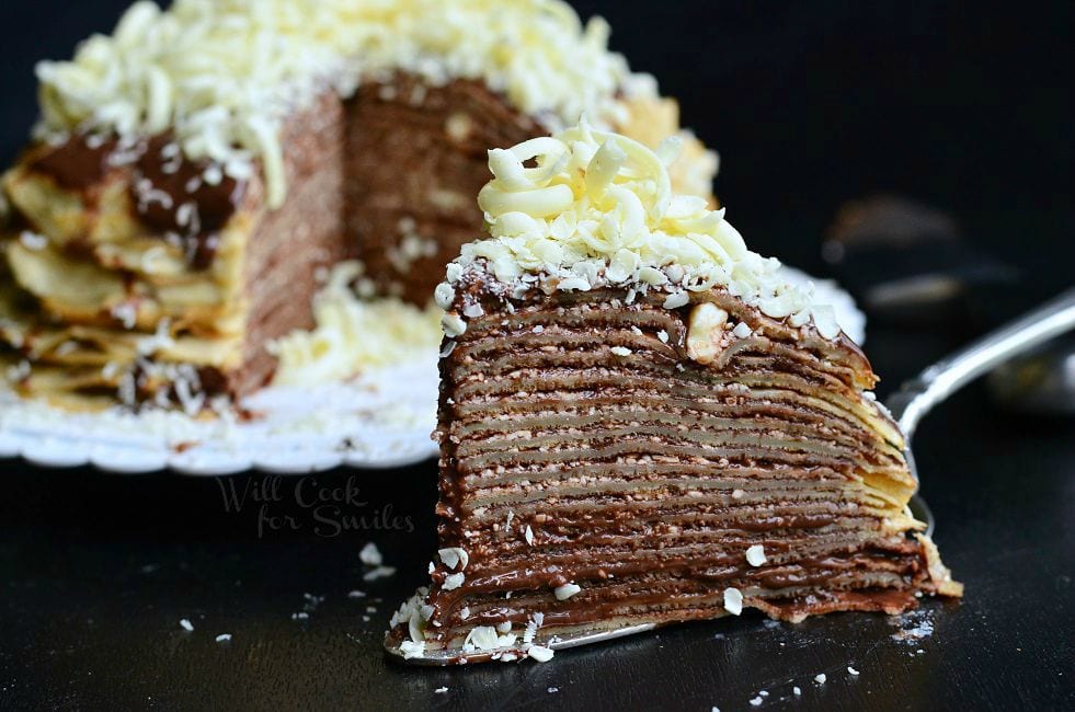 A slice of the Double Chocolate Chocolate Pudding Crepe Cake sits on a serving spatula. The stacked crepes make for thin layers. Chocolate is a key element of this cake. It's between all layers and then on top as well. There are also white chocolate shavings on top of this slice of cake. The rest of the cake fades into the background.