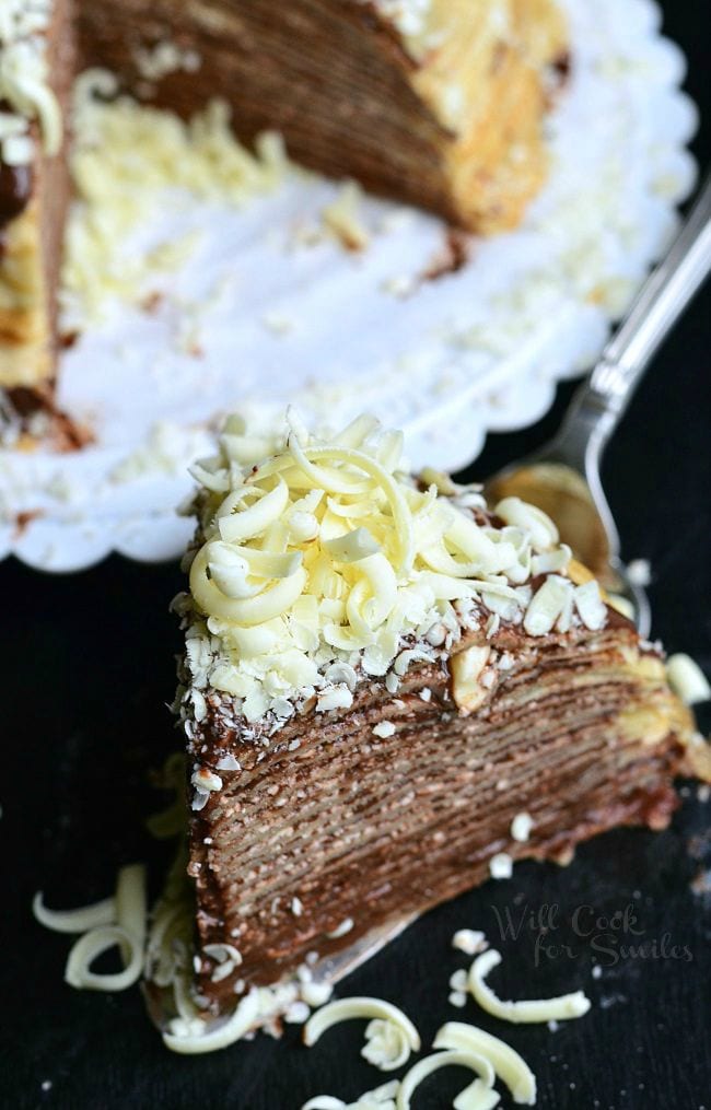 Top view of a slice of the Double Chocolate Chocolate Pudding Crepe Cake sitting on a serving spatula. The stacked crepes make for thin layers. Chocolate is a key element of this cake. It's between all layers and then on top as well. There are also white chocolate shavings on top of this slice of cake.