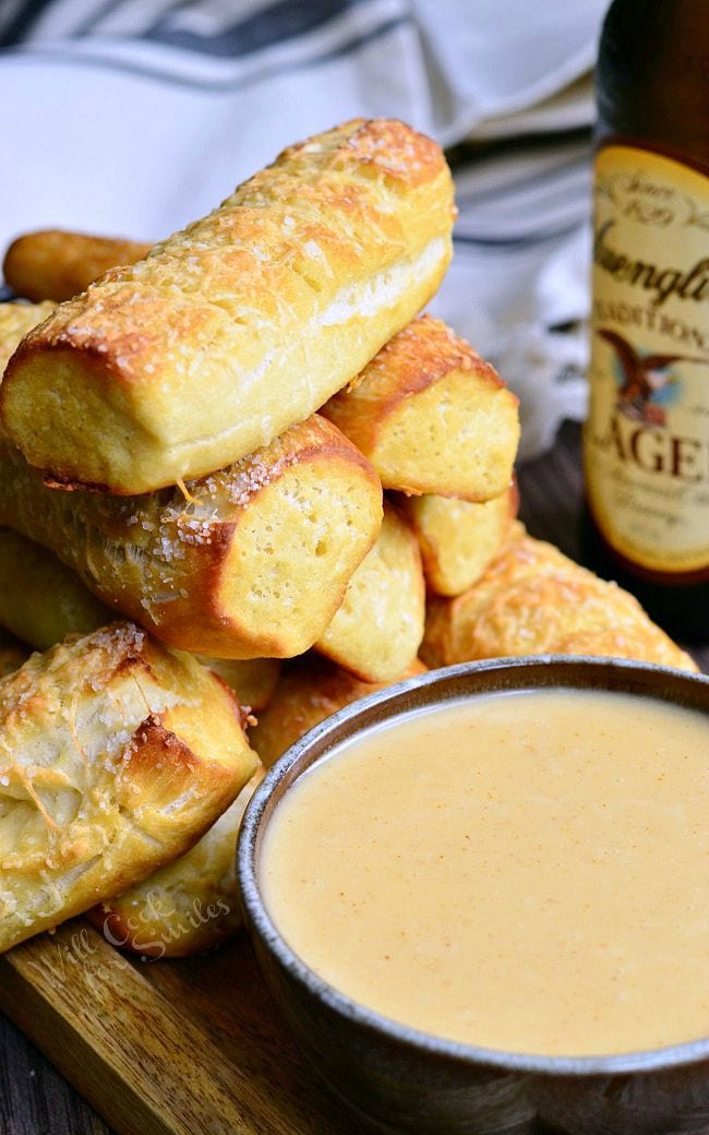 A mound of Homemade Soft Pretzels placed on a wooden board. Pretzels are a nicely golden brown. Infront of the pretzels, is a bowl of beer cheese dip. In the background, there is a bottle of beer.