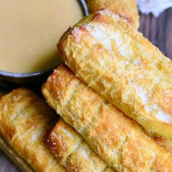 brown bowl filled with beer cheese sauce while homemade parmesan soft pretzel sticks are stacked in the foreground on a wooden cutting board