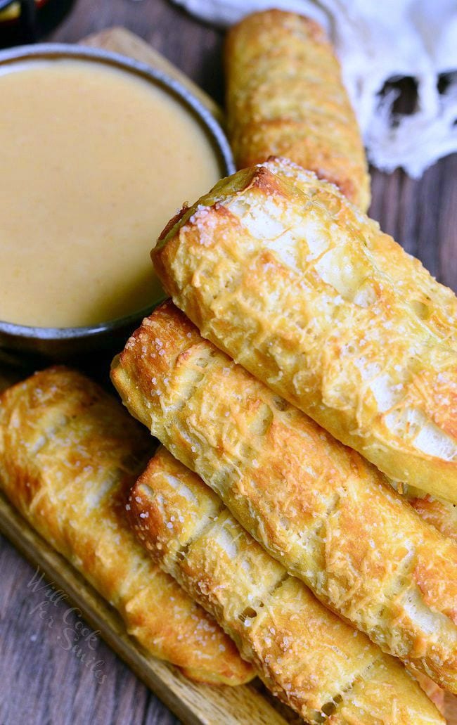 brown bowl filled with beer cheese sauce while homemade parmesan soft pretzel sticks are stacked in the foreground on a wooden cutting board