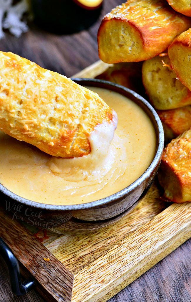 One Homemade Soft Pretzel being dipped into the bowl of beer cheese dip. To the right is other pretzels piled on the wooden board.