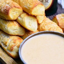 brown bowl filled with beer cheese sauce while homemade parmesan soft pretzel sticks are stacked in the background on a wooden cutting board and a bottle of Yuengling stands in the back right