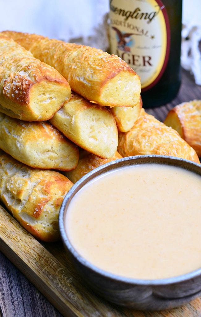 A mound of Homemade Soft Pretzels placed on a wooden board. Pretzels are a nicely golden brown. Infront of the pretzels, is a bowl of beer cheese dip. In the background, there is a bottle of beer.