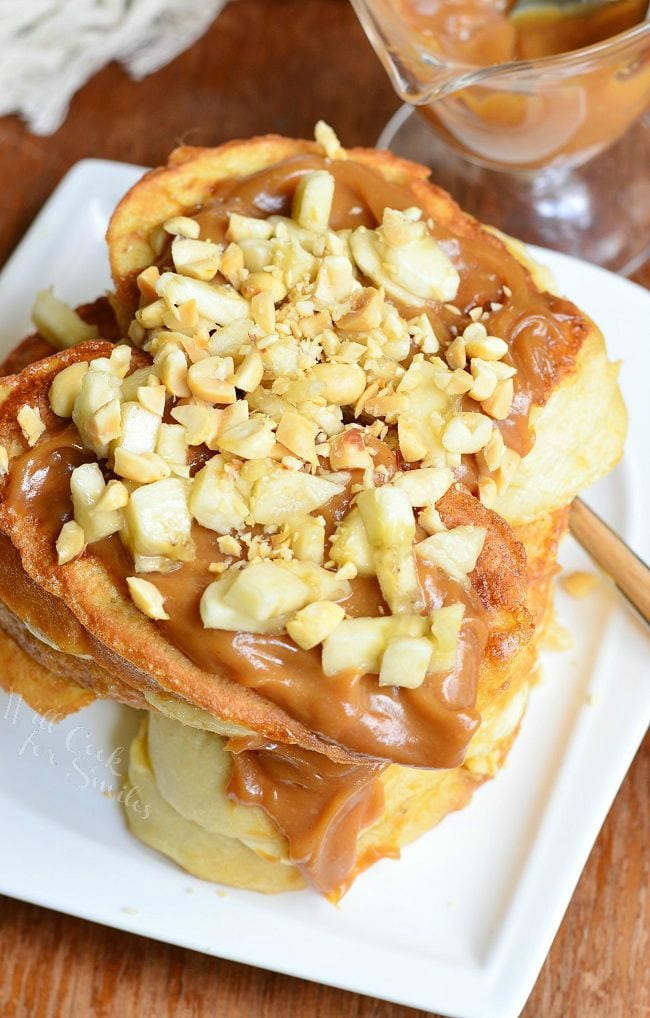 Top view of Stacked Peanut Butter Banana Stuffed French Toast served on a white dish. Peanut butter sauce is smothered on top. Chopped peanuts and banana pieces are also on top.