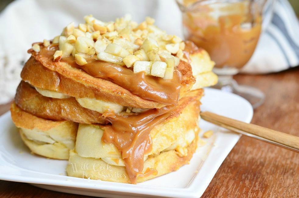 Stacked Peanut Butter Banana Stuffed French Toast served on a white dish. Peanut butter sauce is smothered on top and goes down the side. Chopped peanuts and banana pieces are also on top.