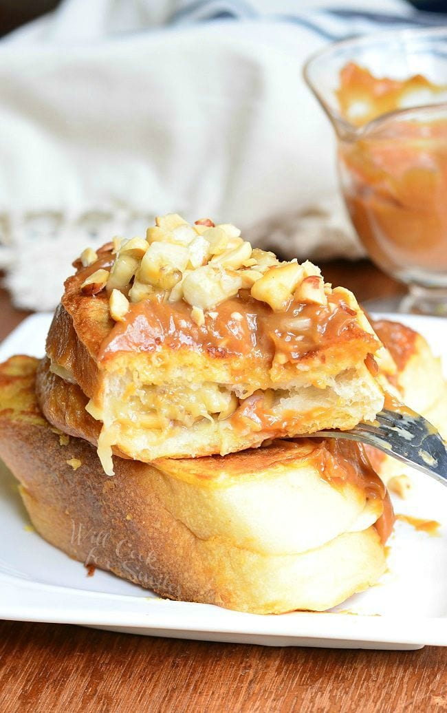 Stacked Peanut Butter Banana Stuffed French Toast served on a white dish. Top piece is cut to show the filling. Peanut butter sauce is smothered on top with chopped peanuts and banana pieces.