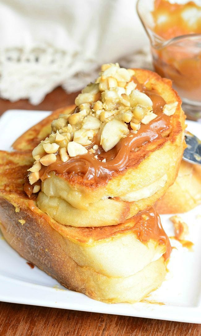 Stacked Peanut Butter Banana Stuffed French Toast served on a white dish. Peanut butter sauce is smothered on top. Chopped peanuts and banana pieces are also on top.