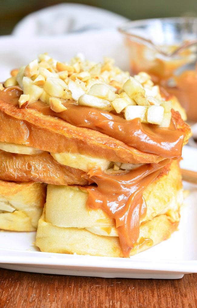 Stacked Peanut Butter Banana Stuffed French Toast served on a white dish. Peanut butter sauce is smothered on top and goes down the side. Chopped peanuts and banana pieces are also on top.