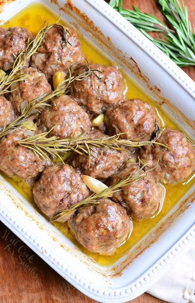 Top view of Rosemary Garlic Baked Meatballs in a white baking dish with rosemary and pieces of garlic after being cooked.
