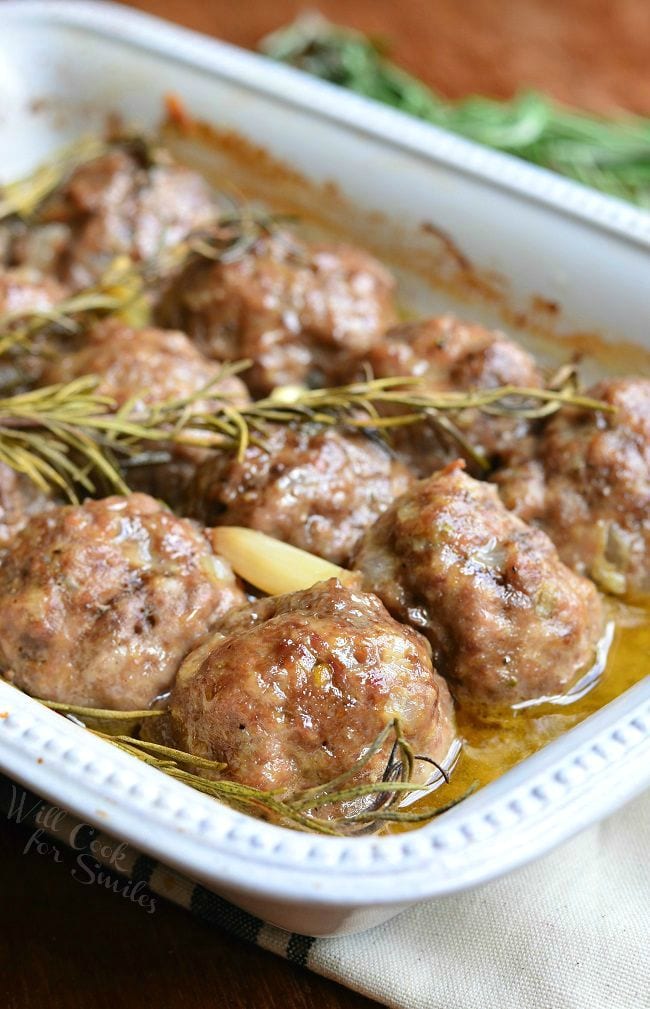 Rosemary Garlic Baked Meatballs in a white baking dish with rosemary and pieces of garlic after being cooked.