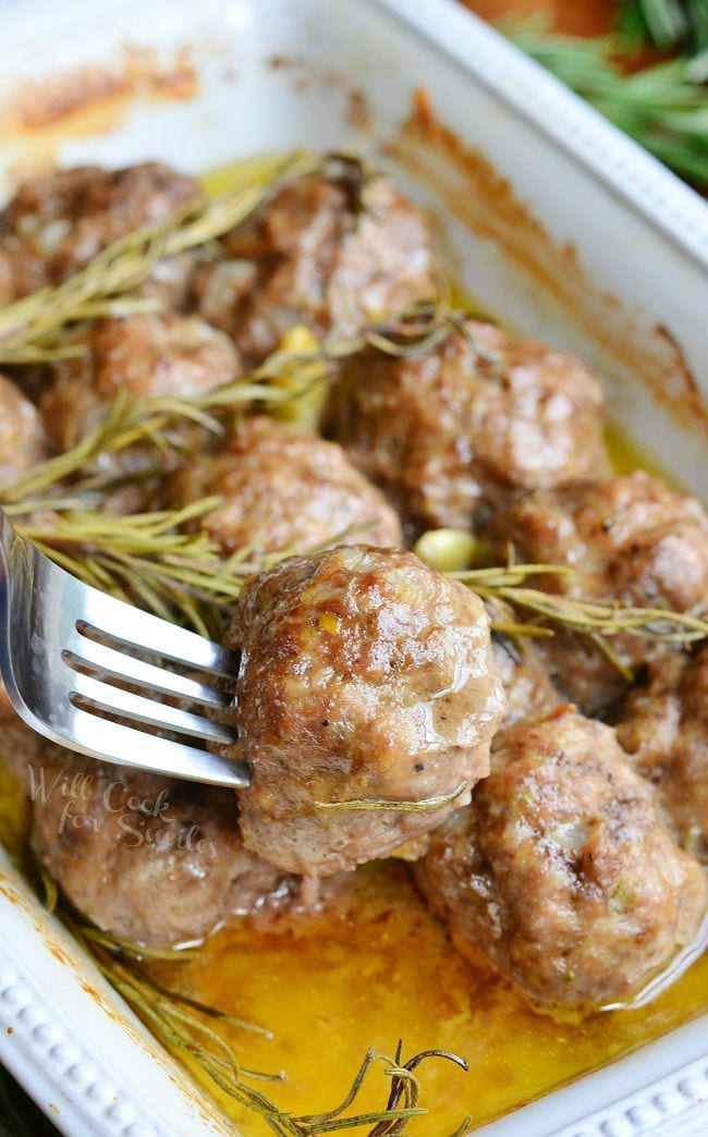 Rosemary Garlic Baked Meatballs in a white baking dish with rosemary and pieces of garlic after being cooked. A fork is inserted into one meatball holding it above the others.