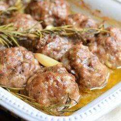 close up view of roasted garlic rosemary baked meatballs in a white baking dish on a wooden table topped with 3 rosemary sprigs