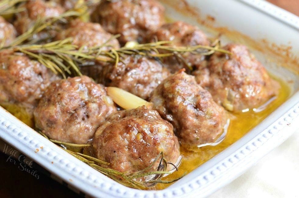 Rosemary Garlic Baked Meatballs in a white baking dish with rosemary and pieces of garlic after being cooked.