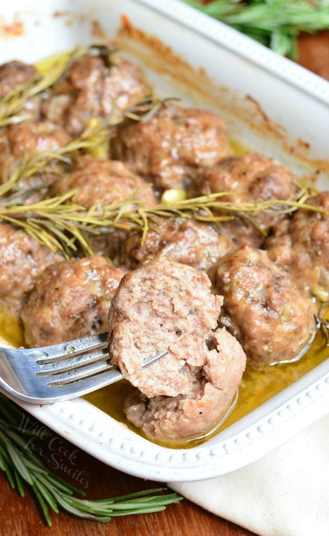 Rosemary Garlic Baked Meatballs in a white baking dish with rosemary and pieces of garlic after being cooked. A fork is holding up half a meatball above the dish.