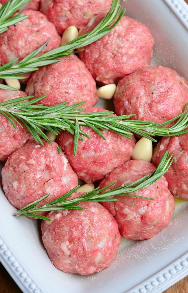 Uncooked meatballs in a white baking dish with rosemary and pieces of garlic.