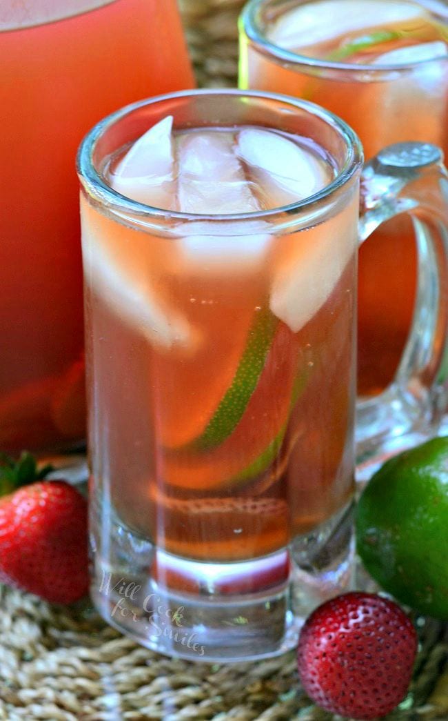 Strawberry Lime Infused Iced Tea is presented in a glass mug. There is floating ice, as well as strawberry and lime slices in the glass. An additional mug and pitcher are in the background. A couple strawberries and lime lay on the table.